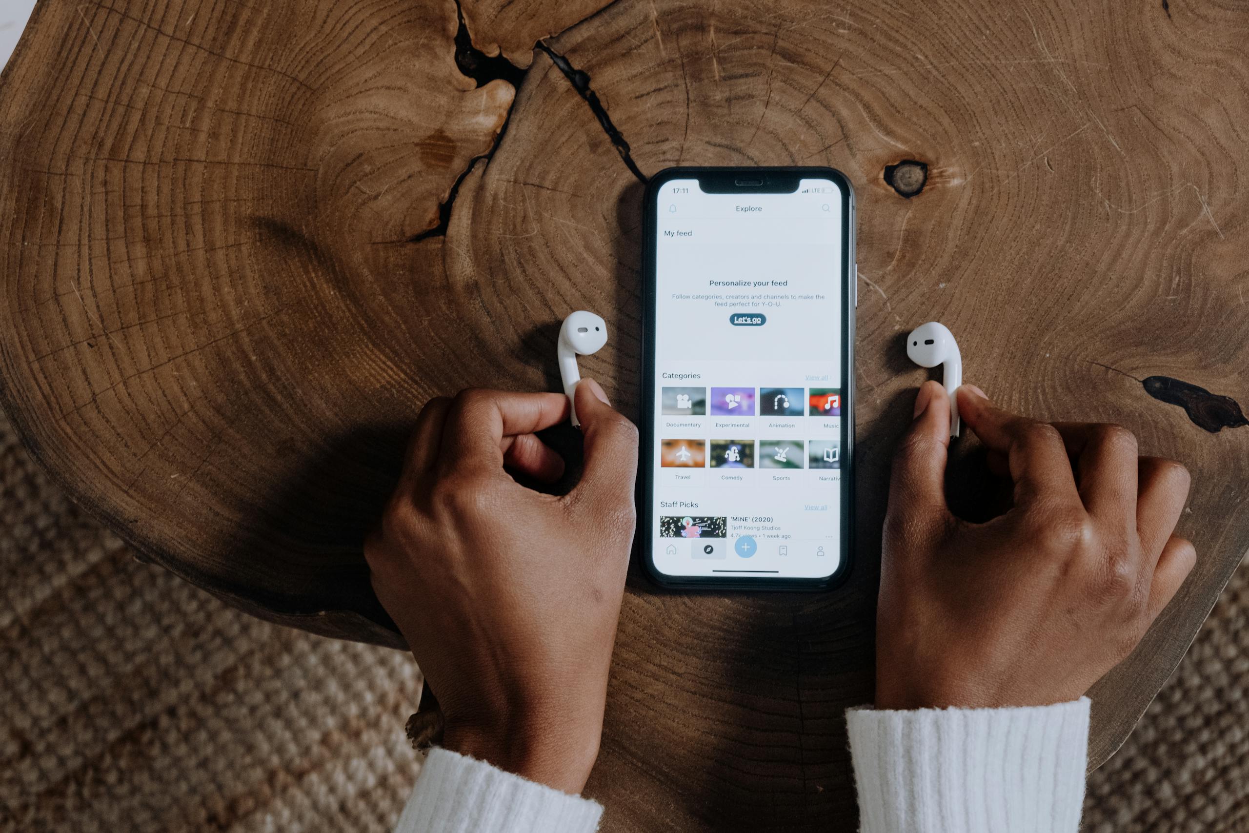 Hands holding wireless AirPods next to a smartphone displaying a social app on a wooden surface.